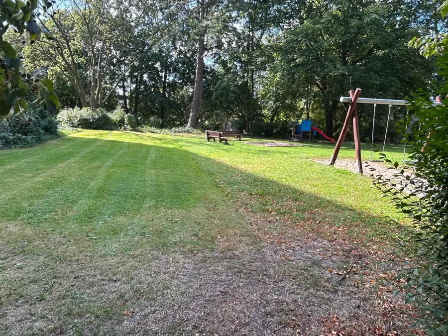 Frisch gemähte Rasenfläche mit hellen und dunklen Streifen, rechts eine Holzschaukel, dahinter Bäume und Bänke